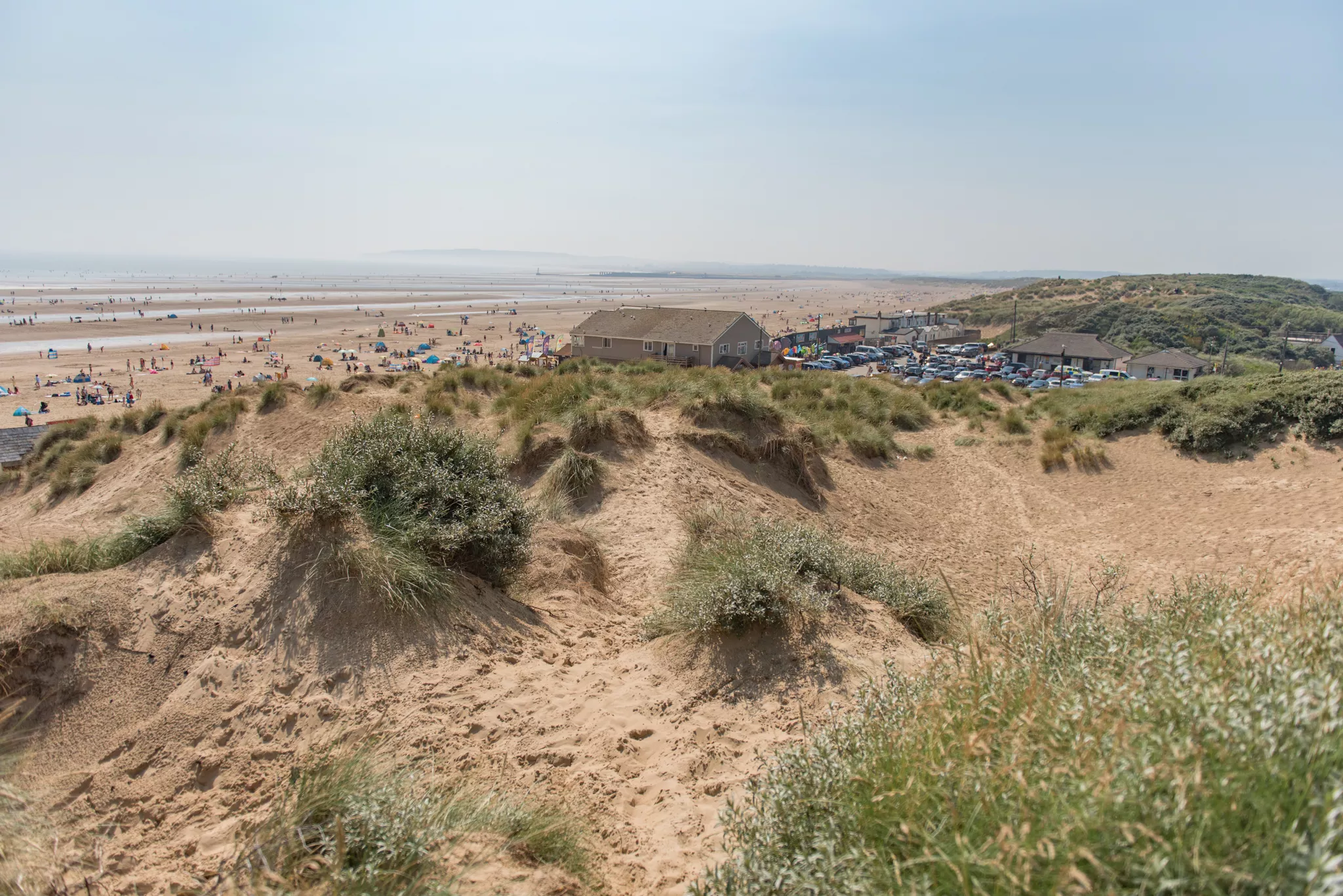 A photo of the sand dunes at Camber Sands beach in Sussex, there's lots of people spread out across the sea front and in the water.