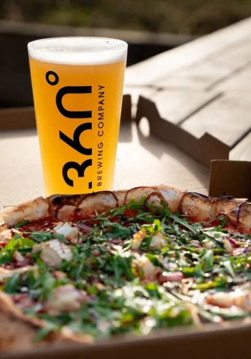 A pint of beer sits behind a freshly-made pizza topped with tomato sauce, brie, bacon and rocket leaves