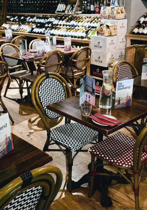 Interiors of Frederic Bistro, Maidstone – a standard bistro layout with tables for two and four, each with patterned bistro-style seating at either side of the table. There are shelves filled with bottles of fine wines in the background.