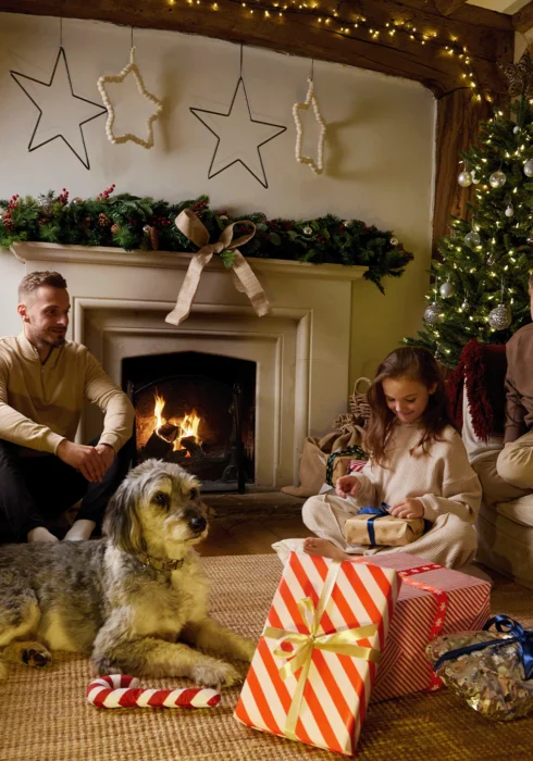 Christmas day at Dormestone Farm, a Bloom Stays holiday home in Kent, where kids are unwrapping presents in front of the fire with a decorate Christmas tree and a dog beside them.