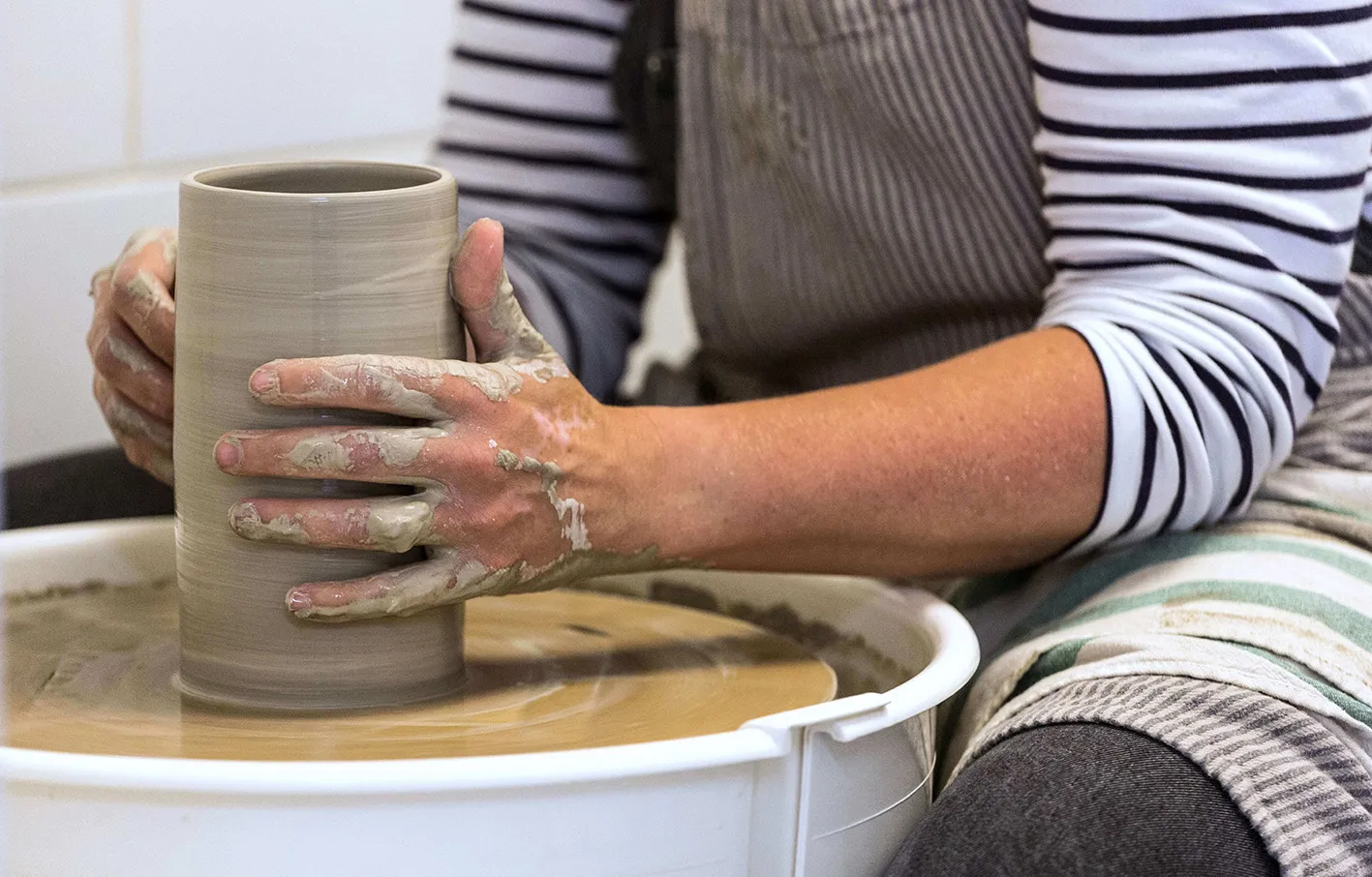 A woman in a striped t-shirt and apron, making a vase; Katharine Gray Ceramics